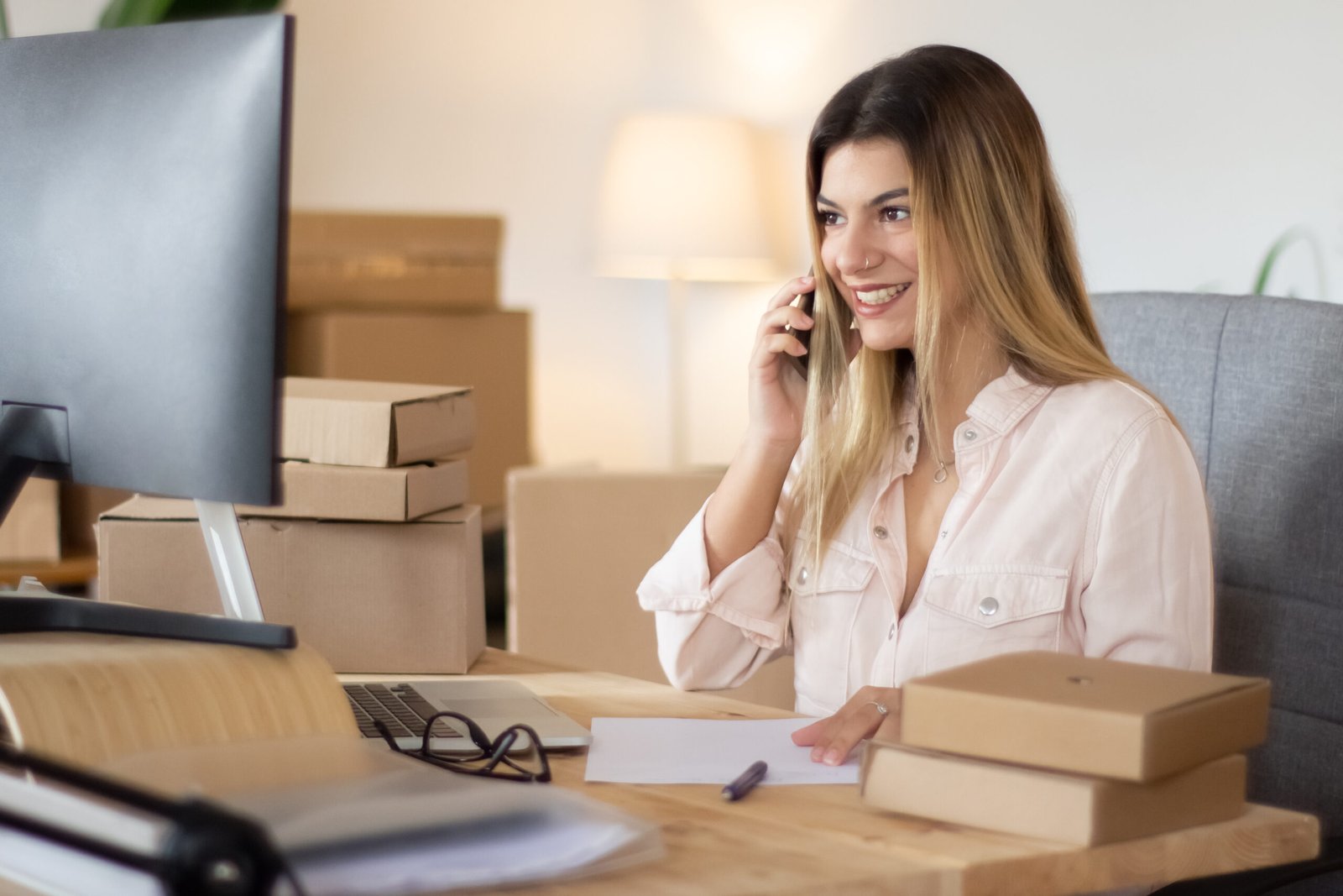 Home Young Online Shop Assistant Consulting Client Phone Looking Computer Screen Smiling Beautiful Caucasian Woman Working Home Office Surrounded By Cardboard Parcels E Commerce Concept 1 Scaled
